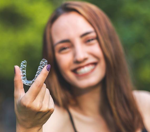 Girl in front of green trees holding out Invisalign smiling